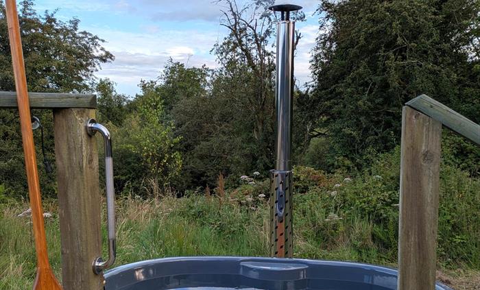 A grey hot tub with chimney, with a wildflower meadow and trees behind